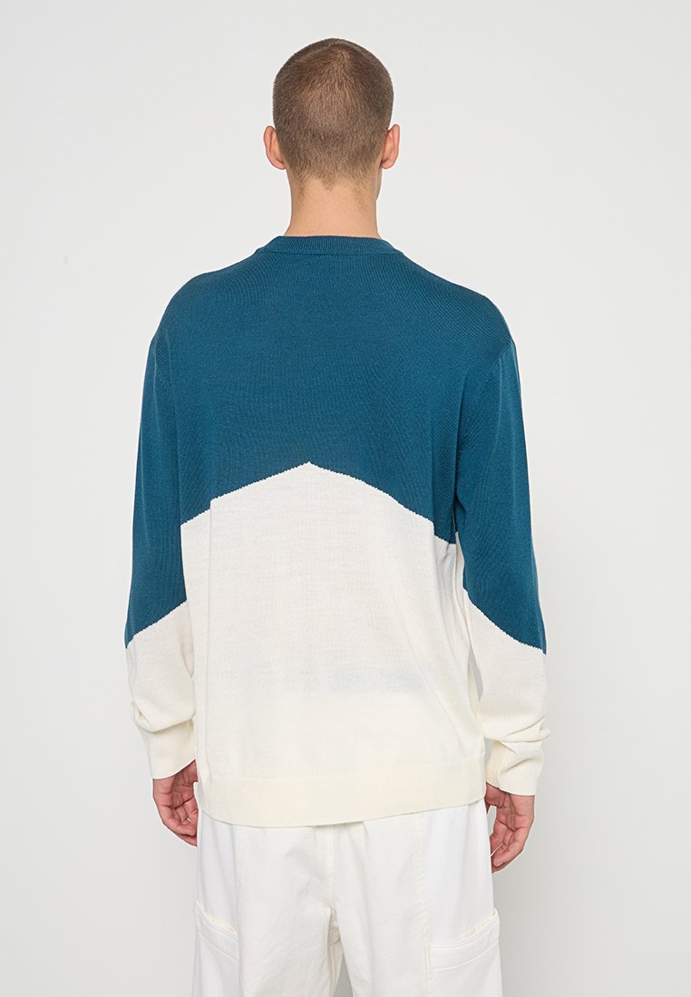 Muškarac nosi džemper s dugim rukavima pleten u tirkiznoj i bijeloj boji s geometrijskim blokovima boja, u kombinaciji s širokim bijelim hlačama, prikazan odostraga.