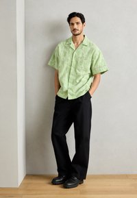 Short-sleeve green shirt with sun patterns, pocket; paired with loose black trousers and black shoes, standing against a light wall.