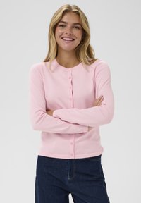 Una donna sorridente con capelli biondi che indossa un cardigan rosa con bottoni e jeans blu scuro, in piedi con le braccia incrociate su uno sfondo chiaro.