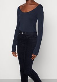 Mörkblå långärmad bodysuit med vid rund halsringning, i kombination med mörka kordbyxor med framfickor och en synlig metallknapp.