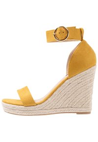 Žluté semišové sandály na wedges s ankletovým páskem a kulatou přezkou, s texturovaným základem omotaným provázkem. Design s otevřenou špičkou.