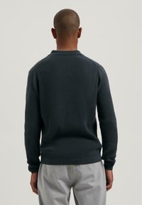 Man with close-cropped hair wearing a dark textured long-sleeve sweater and light gray pants, facing away against a plain background.