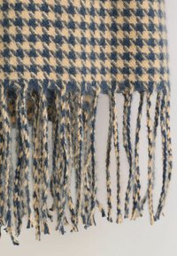 Blå og beige houndstooth mønstret uld tørklæde med snoede frynser hængende mod en lys baggrund.