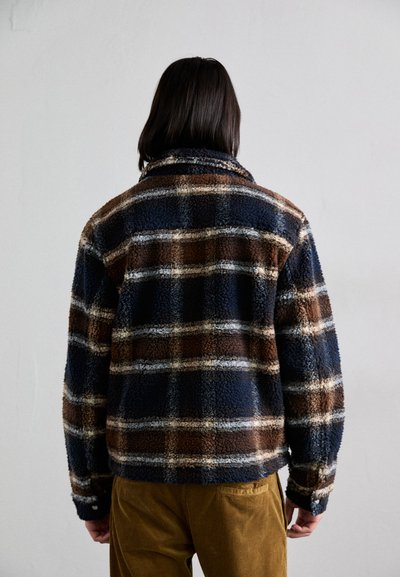 Casaco de fleece com um padrão de xadrez azul, castanho e creme. Textura suave com colarinho, bolsos laterais e punhos com botão.