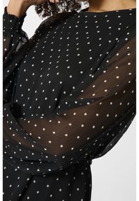 Blouse noire transparente à pois blancs, avec manches longues bouffantes et poignets froncés. Tissu doux et fluide avec une coupe décontractée.