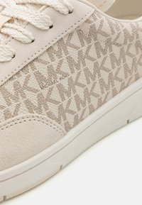 Beige sneaker, texturált, mintás felsőrésszel, amelyen ismétlődő "MK" logó található. Fűzős kialakítás sima, könnyű talppal.