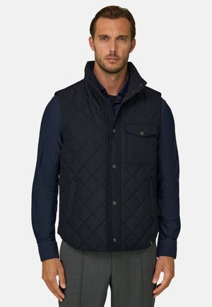 Mann trägt eine schwarze gesteppte Weste über einem dunkelblauen Langarmshirt und graue Hose, steht vor einem einfarbigen hellen Hintergrund.
