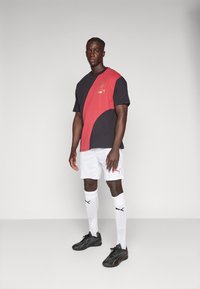 Svart och röd Puma-t-shirt med en böjd design; kombinerad med vita shorts och svarta sportskor; vanliga vita strumpor med logotyp.