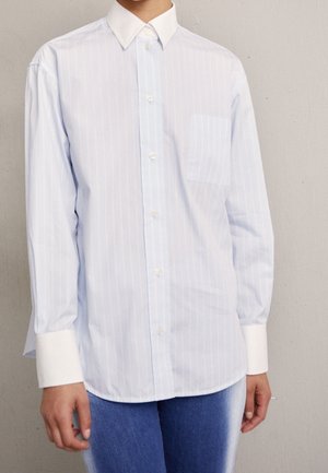 Person iført en lyseblå, stribet skjorte med hvid krave og manchetter, kombineret med blå gradient jeans, står mod en neutral væg.