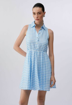 Frau mit dunklen Haaren, die ein ärmelloses blau-weiß kariertes Hemdkleid vor einem schlichten hellen Hintergrund trägt.