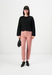 Svart texturerad stickad tröja, rosa uppvikta byxor och svarta snörskor. Modellen håller en liten svart väska. Enkel, neutral bakgrund.