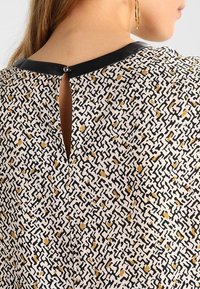 Mönstrad blus med vit botten, med svarta och gyllene abstrakta mönster. Svart halskant och en liten knäppning bak.