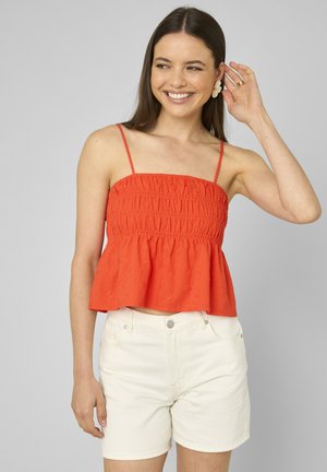Jeune femme souriante, portant un haut rouge sans manches froncé avec bretelles spaghetti et un short blanc taille haute, ajustant une boucle d'oreille en forme de fleur.
