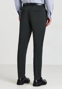 Mann in dunkelgrauen Anzughosen, schwarzen Schuhen und einem hellblauen gestreiften Hemd, der vor einem schlichten hellen Hintergrund steht.