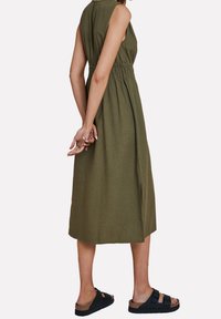 Olijfgroene mouwloze jurk met een strakke taille en split aan de zijkant, gemaakt van lichtgewicht stof. Zwarte sandalen met gespen maken de look compleet.
