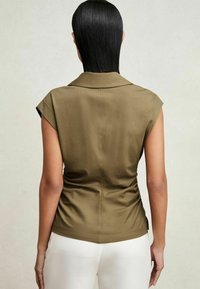 Persona con cabello corto negro que lleva una blusa sin mangas de color verde oliva con cuello y pantalones blancos, mostrada de espaldas contra un fondo neutral.