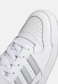 White leather sneaker with perforated toe, light gray accents, and three-stripe design. Features flat laces and rubber sole.