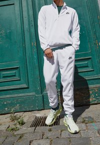 Vit zip-up Nike-jacka med matchande sweatpants, med svarta logotypdetaljer. Bärs med ljusgröna träningsskor, mot en grön bakgrund.