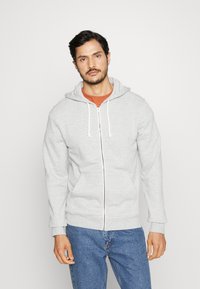 Grijze zip-up hoodie met een trekkoord capuchon en voorkruiszakken, gemaakt van zachte stof. Draagt over een oranje t-shirt, gecombineerd met blauwe jeans.