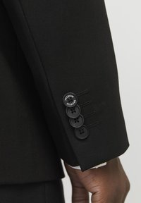 Zwarte colbertmanchet met vier zwarte knopen, waarvan er één de tekst "PREMIUM" heeft, gedetailleerde stiksels en een gladde stoffen textuur.