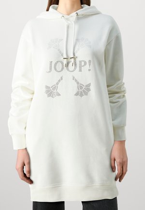 Kobieta ubrana w długi biały hoodie z "JOOP!" i dekoracyjnymi wzorami ze srebrnych ćwieków, stoi z rękami swobodnie opuszczonymi wzdłuż ciała.
