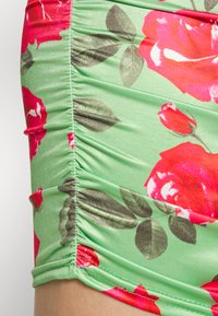 Calções de tecido franzido em verde com um padrão floral apresentando rosas vermelhas e folhas verdes. Textura suave com detalhes em elástico.