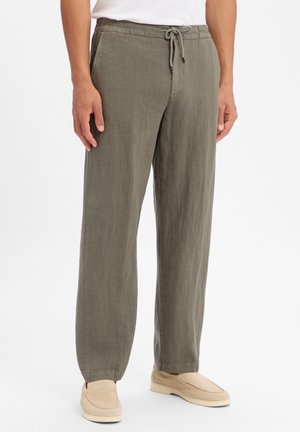 Mann trägt olivgrüne Leinenhose mit Kordelzug und beige Slipper, steht vor weißem Hintergrund.