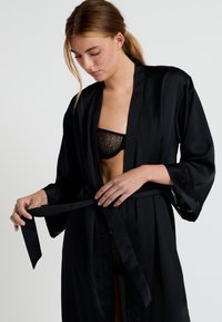 Kimono en satin noir avec des manches larges, noué à la taille. Présente des accents de dentelle délicats sur les bords des manches.