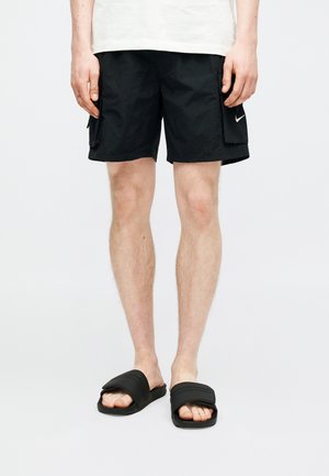 Άτομο που φοράει μαύρα cargo σορτς Nike και μαύρα σανδάλια τύπου slides, στέκεται μπροστά σε ένα απλό ανοιχτόχρωμο φόντο.