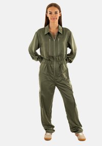 Jumpsuit verde oliva con zip frontale, due tasche sul petto con zip, vita elasticizzata e zip laterali. Tessuto leggero e liscio.