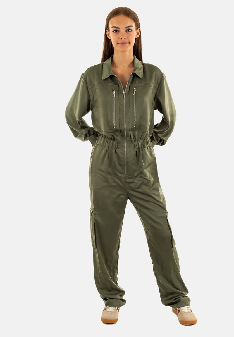 Jumpsuit verde oliva con zip frontale, due tasche sul petto con zip, vita elasticizzata e zip laterali. Tessuto leggero e liscio.