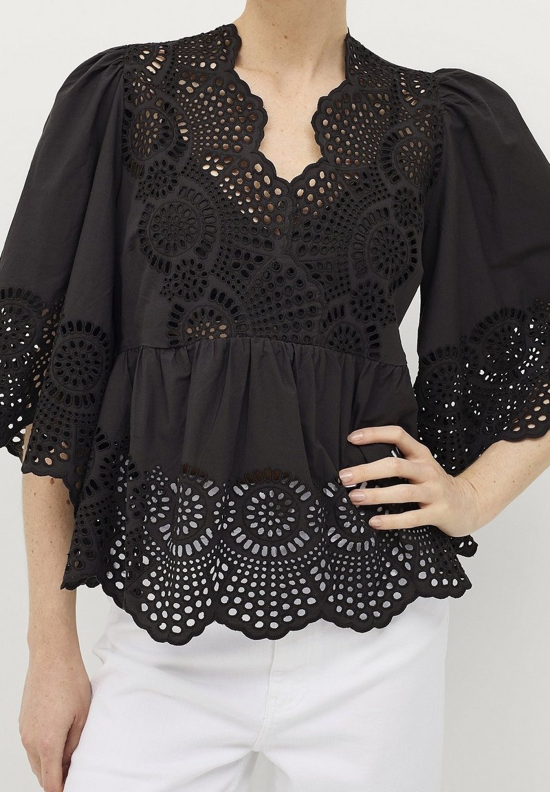 Blouse noire avec des motifs complexes en dentelle ajourée circulaire sur la poitrine, les manches et l'ourlet, associée à un pantalon blanc, main sur la hanche.