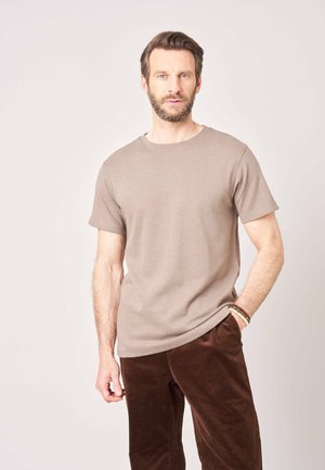 Man met baard, gekleed in een beige shirt met korte mouwen en een bruine broek, staat tegen een effen lichte achtergrond met één hand in de broekzak.