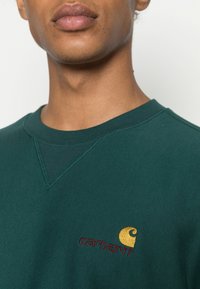 Sweatshirt verde com decote e costuras dos ombros canelados, apresentando um logotipo "Carhartt" em dourado e bordô no peito. Material em algodão.