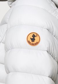 Tissu de doudoune blanche avec texture matelassée. Présente un patch circulaire en tissu orange avec un canard noir et le texte « SAUVEZ LE CANARD ».