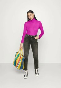 Sieviete, kuri valkā spilgti rozā augsto apkakli, melnus džinsus un melnbaltas sporta kurpes, turēt krāsainu svītrainu rokassomu ar dzeltenām rokturiem.