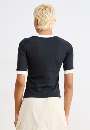 Persoon met kort blond haar, van achteren gezien, draagt een zwart T-shirt met korte mouwen met witte bies en een beige rok met zijzakken.