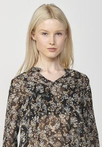 Blusa nera floreale con scollo a V, realizzata in un tessuto trasparente decorato con delicati motivi di fiori bianchi e dorati.