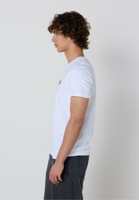 Balta medvilninė marškinėliai su apvalia iškirpte, trumpomis rankovėmis ir mažai spausdinta detale ant krūtinės. Dėvimi su pilkais kelnėmis, rodomi šoniniame profilyje.