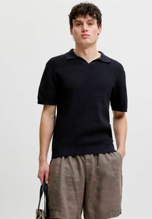 Junger Mann trägt ein schwarzes kurzärmeliges Strick-Poloshirt und lockere braune Shorts, hält eine beige Tragetasche mit schwarzen Griffen.