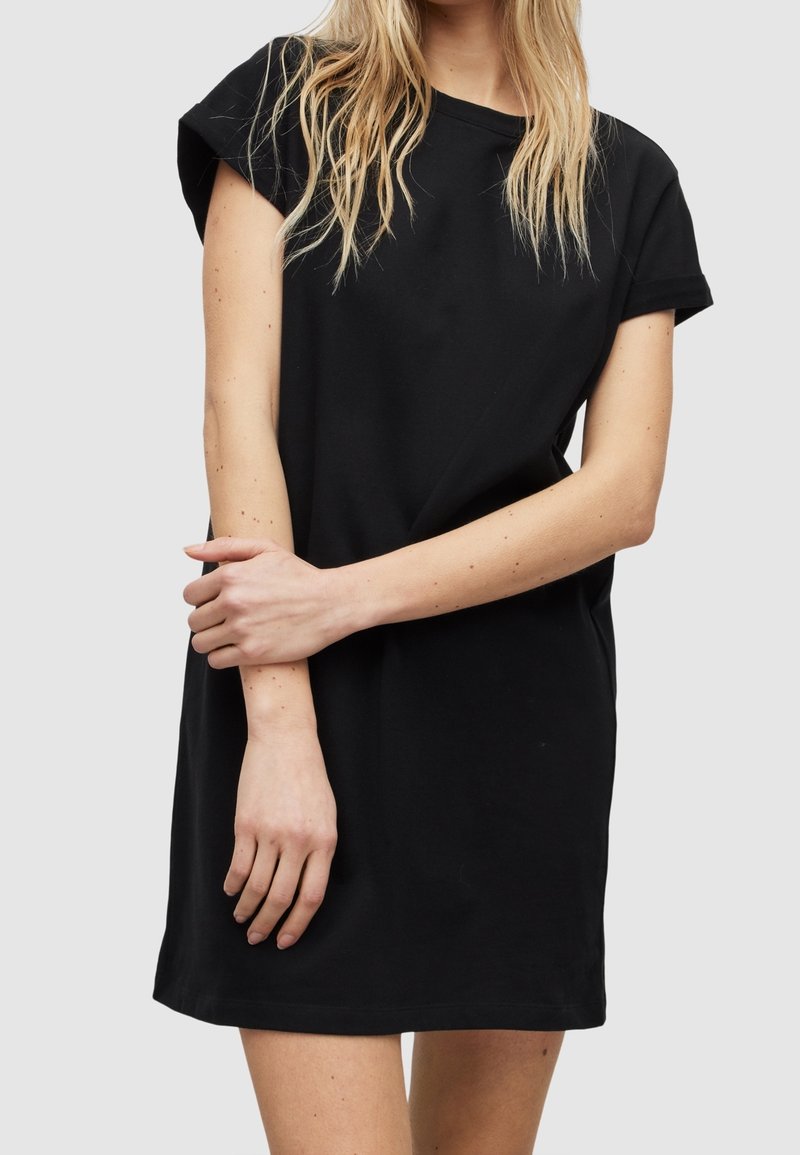 Femme aux longs cheveux blonds portant une robe noire à manches courtes, debout, un bras croisé sur l'autre, devant un fond uni.