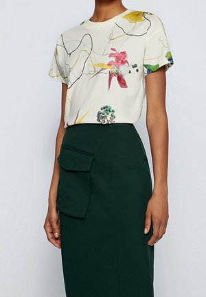 Kvinde iført en kortærmet hvid bluse med farverige blomsterprint og en mørkegrøn nederdel med høj talje og en stor lomme foran.