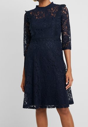 Robe en dentelle bleu marine avec manches trois-quarts, col montant et motifs floraux, cintrée à la taille avec une bordure décorative.