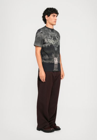 Male model with curly hair stands facing right wearing a black graphic t-shirt, loose dark pants, and dark shoes against a plain background.
