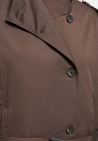 Gros plan de l'avant d'un manteau marron montrant les boutons, les boutonnières et les détails de tissu superposés sur le col et le corps.