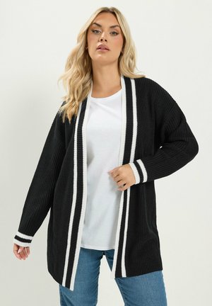Kobieta o blond włosach nosi czarny otwarty kardigan z białym, prążkowanym wykończeniem, białą koszulę i niebieskie jeansy na jednolitym tle.