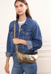 Sac à main en métal doré avec une forme incurvée et une texture lisse et brillante, tenu par une personne portant une veste en denim et un haut rayé.