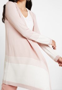 Ljusrosa lång cardigan med krämfärgade ränder, gjord av mjukt tyg. Öppen framtidsdesign, med långa ärmar och en avslappnad passform.