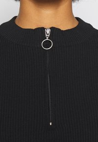 Haut en tissu côtelé noir avec un col montant, doté d'une fermeture éclair avant avec un curseur circulaire et des accents en argent.