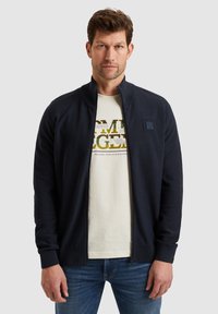 Mornarska zip-up jakna z dvignjenim ovratnikom, teksturirano blago, rebrasti manšeti in sprednjim žepom; nosi se čez svetlo obarvano grafično majico.
